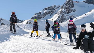 Eine Gruppe von Kindern in Skianzügen steht auf einer schneebedeckten Piste. Im Hintergrund sieht man schneebedeckte Berge und einen klaren blauen Himmel.
