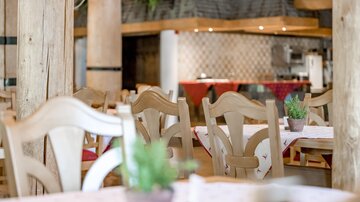An inviting restaurant with wooden chairs and set tables. In the background, you can see a cozy kitchenette.