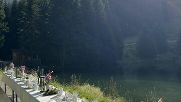 Ein festlich gedeckter Tisch am Ufer eines Teiches, umgeben von hohen Tannen. Die Sonne strahlt über die Landschaft und erzeugt eine angenehme Atmosphäre.
