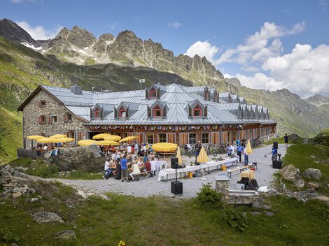Eine charmante Berghütte mit Terrassen und Sonnenschirmen. Im Hintergrund sind majestätische Berge und eine grüne Wiese zu sehen.