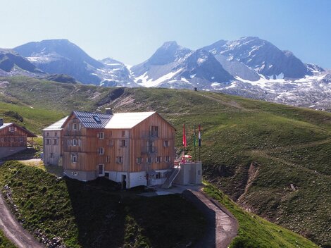 Ein modernes Holzgebäude liegt malerisch in den Bergen. Im Hintergrund sind schneebedeckte Gipfel und eine grüne Landschaft zu sehen.
