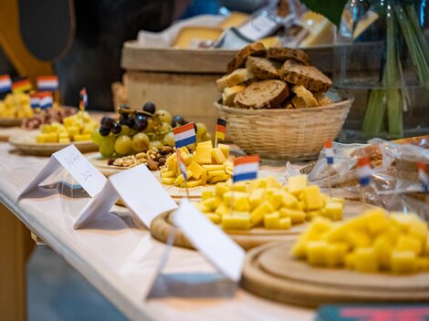 Eine schön dekorierte Käseplatte mit verschiedenen Käsesorten und frischem Brot. Auf dem Tisch sind kleine Flaggen und Obst zu sehen.