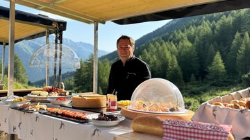 Ein Mann steht an einem Buffet im Freien, umgeben von einer malerischen Berglandschaft. Das Buffet ist mit verschiedenen Speisen und Getränken gedeckt.