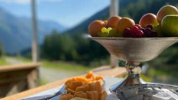 Ein schön angerichteter Tisch mit verschiedenen Käsesorten und einer Schale mit frischem Obst. Im Hintergrund sind Berge und ein blauer Himmel zu sehen.