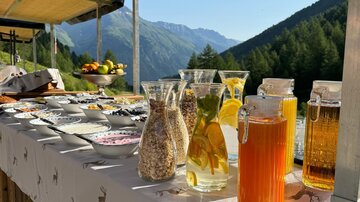 Ein schön gedeckter Tisch im Freien mit verschiedenen Getränken und Speisen. Im Hintergrund sind Berge und ein klarer blauer Himmel sichtbar.