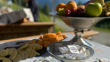 Ein Anrichtetisch mit verschiedenen Snacks, darunter Früchte, Käse und Schokoladen. Im Hintergrund sind Berge und ein blauer Himmel zu sehen.