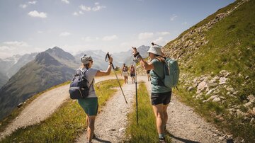 Eine Gruppe von Wanderern geht auf einem Wanderweg in den Bergen. Die Landschaft ist grün mit Bergen im Hintergrund und einem klaren Himmel.