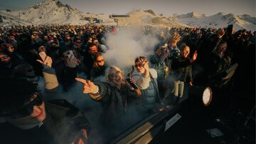 Eine lebhafte Menschenmenge auf einem Event in den Bergen. Rauch und Schnee umgeben die feiernden Personen, die Spaß haben und ihre Smartphones nutzen.