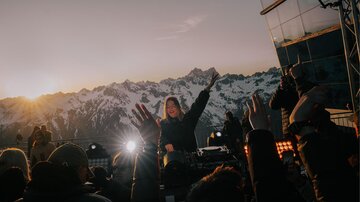 Eine DJane spielt Musik auf einer Bühne mit atemberaubendem Blick auf schneebedeckte Berge. Die Menge tanzt und feiert im Sonnenuntergang.
