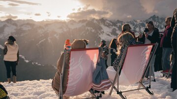 Zwei Personen sitzen entspannt in Liegestühlen auf dem Schnee und genießen den Ausblick auf die Berge. Im Hintergrund sind andere Menschen zu sehen, die die Aussicht ebenfalls schätzen.