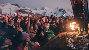 Eine begeisterte Menschenmenge tanzt auf einem Festival in den Bergen. Im Hintergrund sind schneebedeckte Gipfel und ein stimmungsvoller Sonnenuntergang zu sehen.