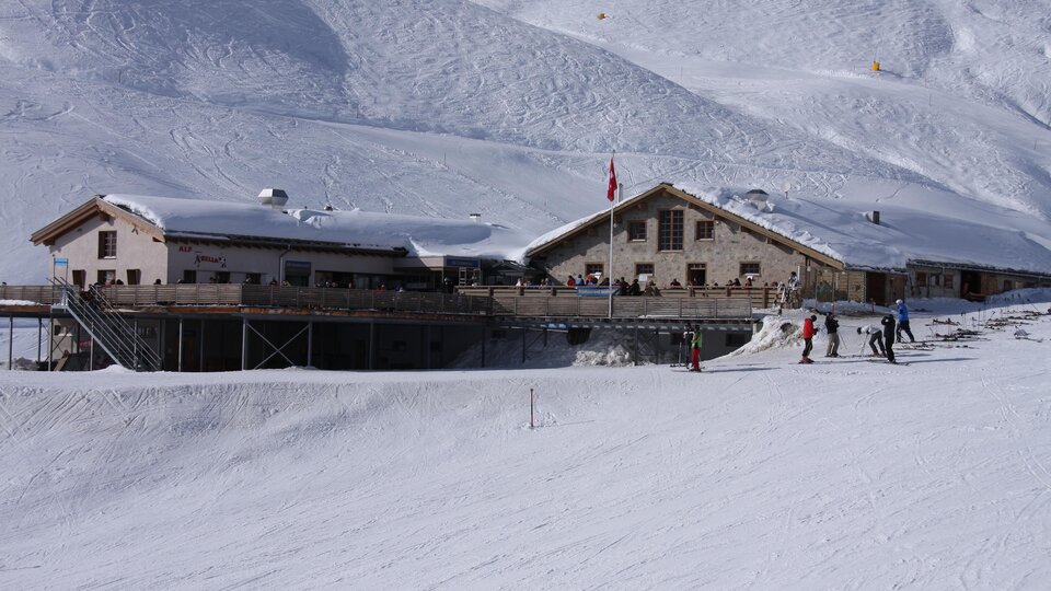 Eine Berghütte im Schnee mit skifahrenden Personen. Die Landschaft ist von schneebedeckten Bergen umgeben.