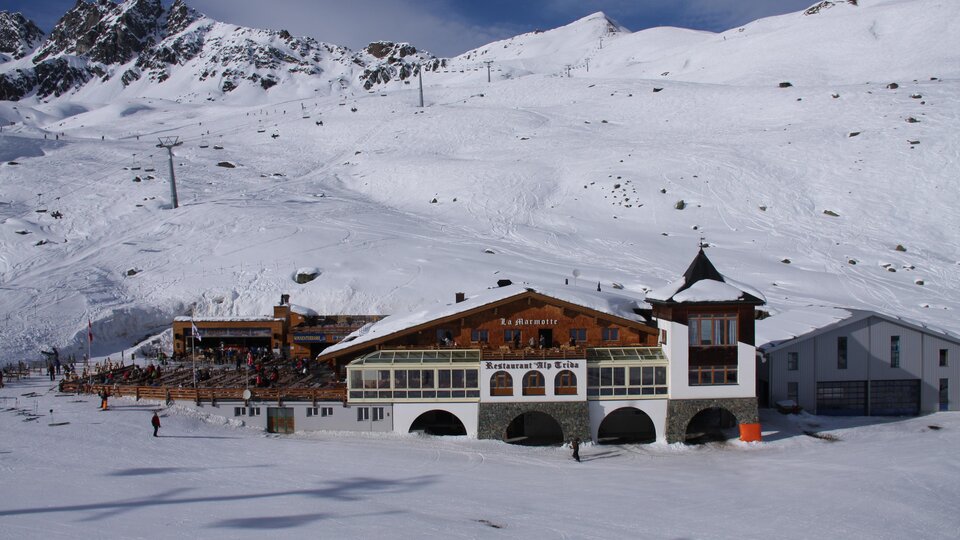 Eine schneebedeckte Landschaft mit Bergmassiven im Hintergrund. Im Vordergrund steht ein traditionelles Berghaus.