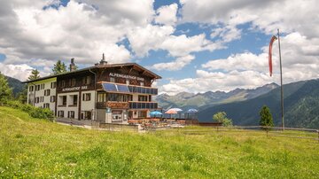 Eine malerische Berghütte umgeben von grünen Wiesen und Bergen. Der Himmel ist teilweise bewölkt und die Atmosphäre ist einladend.