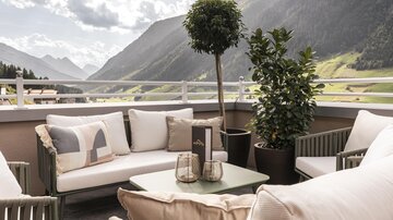 Eine gemütliche Terrasse mit modernen Sitzmöbeln und Pflanzen. Im Hintergrund sind majestätische Berge und ein klarer Himmel zu sehen.