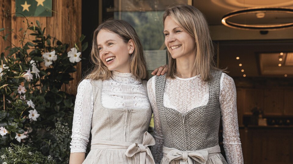 Zwei Frauen stehen lächelnd vor einem Gebäude. Beide tragen traditionelle Kleider mit modernen Akzenten.