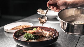 Ein Koch serviert ein Gericht in einer eleganten Schüssel. Die Präsentation umfasst Garnelen, Gemüse und eine Sauce.