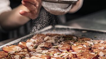 Eine Hand siftet Zucker über ein Blech mit süßen, belegten Platten. Die Oberseite ist mit Mandeln und anderen Zutaten geschmückt.