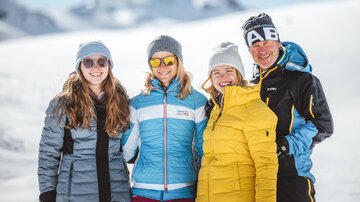 Eine Gruppe von vier Personen steht in einem verschneiten Berggebiet. Alle tragen warme Winterjacken und Mützen, und die Sonne scheint hell.