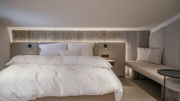 Ein modernes Schlafzimmer mit einem großen Bett und weißen Bettwaren. Eine gemütliche Sitzecke und sanfte Beleuchtung schaffen eine entspannte Atmosphäre.
