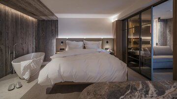 Ein modernes Schlafzimmer mit einem großen Bett und elegantem Holzdesign. Ein freistehender Badebereich und große Fenster sorgen für angenehmes Licht.
