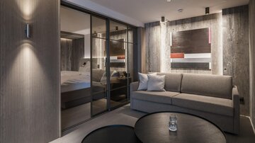 Ein modernes Zimmer mit grauen Wänden und einem komfortablen Sofa. Im Hintergrund ist ein elegantes Schlafzimmer sichtbar.