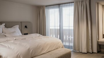 Ein modernes Schlafzimmer mit einem großen Bett und viel natürlichem Licht. Große Fenster mit Vorhängen bieten einen Blick nach draußen.