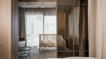 Ein modernes Zimmer mit Holzverkleidung und großen Fenstern. Im Hintergrund steht ein Babybett und der Raum wirkt hell und einladend.