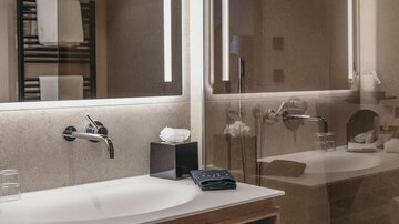 Ein modernes Badezimmer mit einem Waschbecken und großen Spiegeln. Auf dem Regal liegen frische Handtücher und Pflegeprodukte.