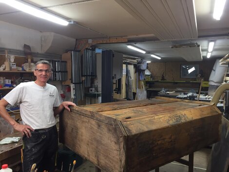 A craftsman stands in his workshop next to a large, old wooden box. Various tools and machines can be seen in the background.