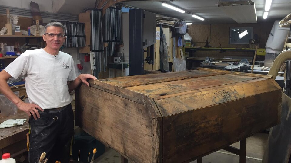 A craftsman stands in his workshop next to a large, old wooden box. Various tools and machines can be seen in the background.