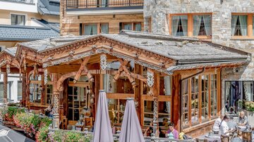Ein gemütliches Restaurant mit Holzverzierungen und Terrassenmöbeln. Umgeben von Blumen und einem malerischen Bergpanorama.