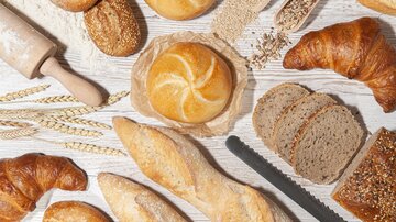 Eine Auswahl an frischem Brot, Brötchen und Croissants liegt auf einer hellen Holzoberfläche. Dazu sind Weizenkörner und ein Nudelholz zu sehen. | © MPREIS Warenvertriebs GmbH