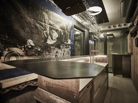 A modern interior with a generous bar counter and rustic wooden elements. On the wall hangs a large black-and-white image depicting a mining scene.