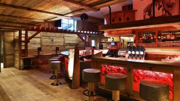 Eine gemütliche Bar mit Holzdekor und warmem Licht. Die Theke ist gut bestückt und es gibt Barhockern zum Sitzen.