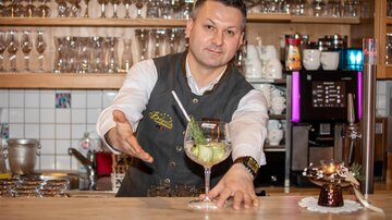 Ein Bartender präsentiert stolz einen farbenfrohen Cocktail an der Bar. Im Hintergrund sind zahlreiche Gläser und eine gut ausgestattete Bar sichtbar.