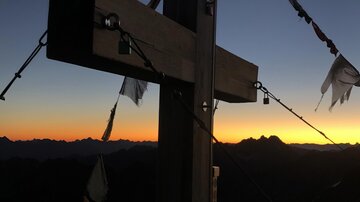 Ein Holzkreuz am Berggipfel, silhouettiert gegen den Sonnenuntergang. Bunte Fahnen wehen im Wind.