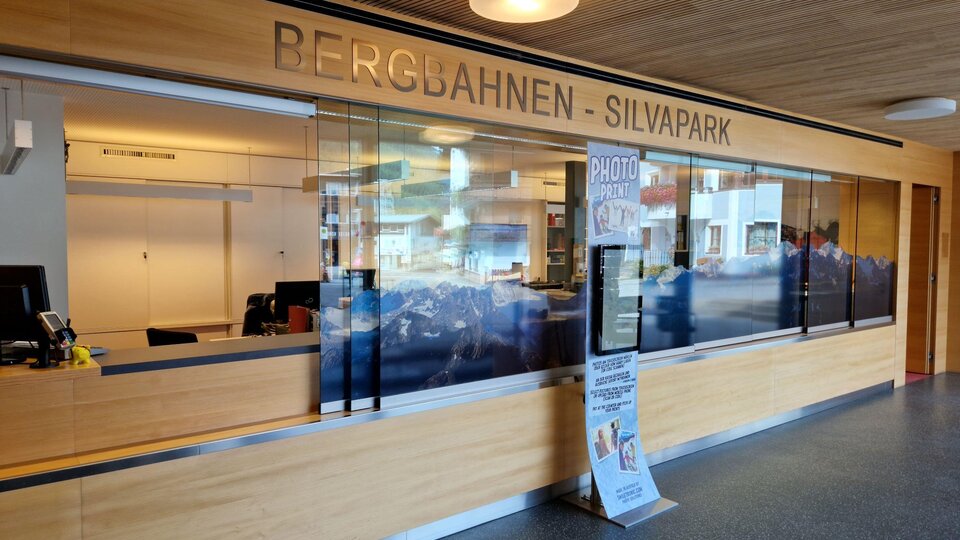 A reception area with wooden paneling and a large glass window. On the wall it says "BERGBAHNEN - SILVAPARK".