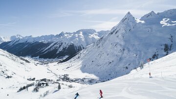 A snow-covered mountain landscape with high peaks and a clear blue sky. Two skiers are skiing down the slope.