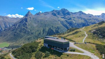 A modern building in the mountains, surrounded by green meadows and tall peaks. The sky is clear and blue, perfect for outdoor activities.
