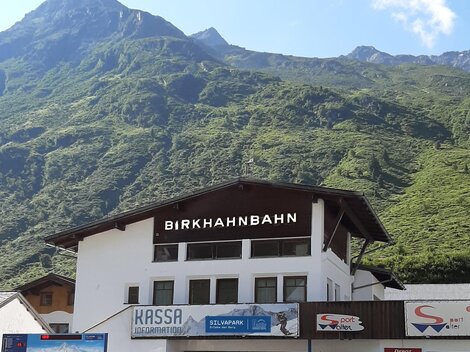A mountain cable car station named "Birkhahnbahn" is located in the foreground. In the background, green mountains and a blue sky can be seen.