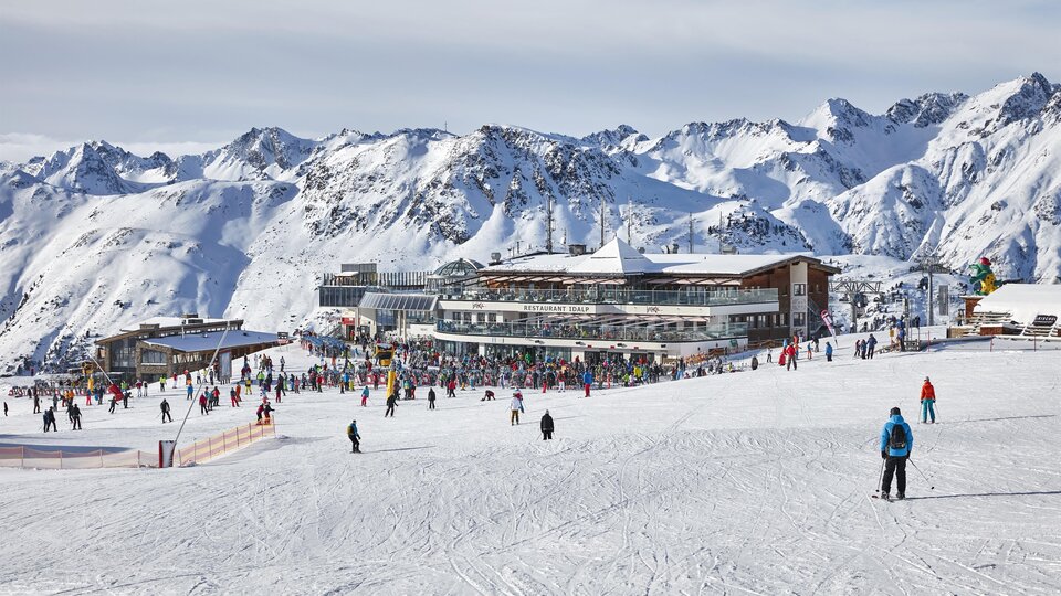 Ein belebtes Skigebiet mit vielen Menschen auf der Piste. Im Hintergrund sind schneebedeckte Berge und ein modernes Gebäude zu sehen.