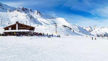 Ein schneebedeckter Berg mit einer Skihütte und vielen Menschen. Im Hintergrund sind majestätische Berge unter einem klaren Himmel zu sehen.