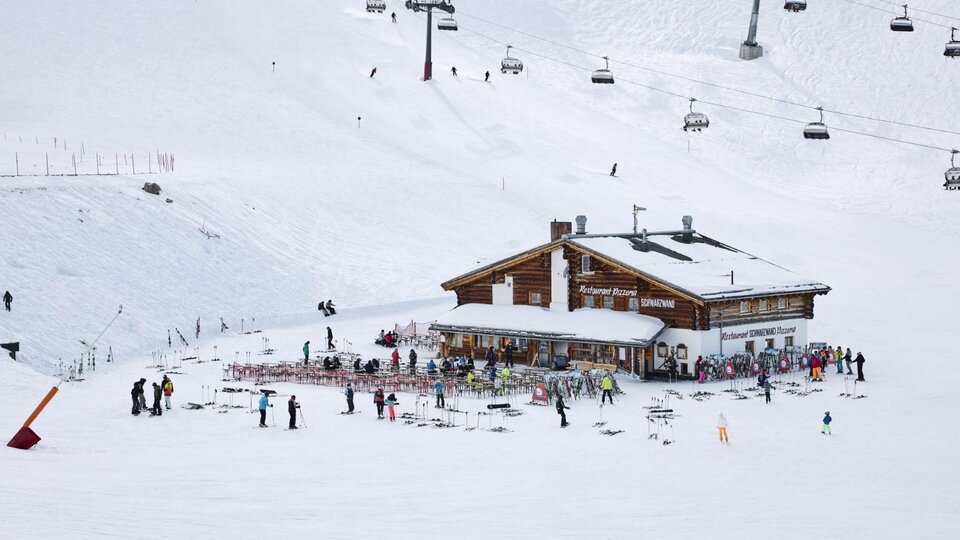 Ein Holzhaus in den Bergen, umgeben von Schnee und Skifahrern. Im Hintergrund sind Skilifte zu sehen.