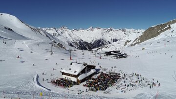 Eine schneebedeckte Berglandschaft mit einem Skihütte im Vordergrund. Viele Skifahrer sind auf den Pisten unterwegs, und die Gipfel sind klar und sonnig.