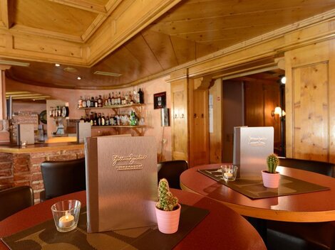 A cozy restaurant with wooden walls and an inviting bar. Menu cards and small potted plants are on the tables.