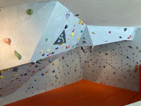 Eine Kletterwand in einem Innenraum mit vielen bunten Griffen. Der Boden ist mit einer orangen Matte ausgelegt.