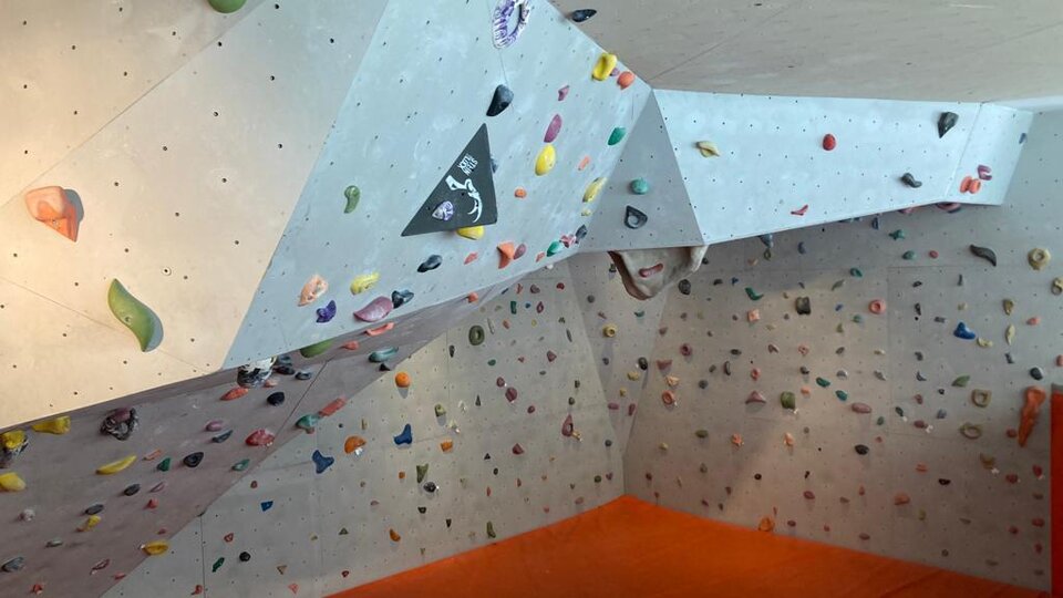 Eine Kletterwand in einem Innenraum mit vielen bunten Griffen. Der Boden ist mit einer orangen Matte ausgelegt.