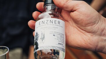 A handheld bottle with the label "ENZNER" is in the foreground. In the background, blurred people and a wooden bench can be seen.