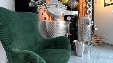 A cozy green armchair is positioned in front of a shiny copper and stainless steel distillation setup. The room has a modern ambiance with a clear, minimalist decor.